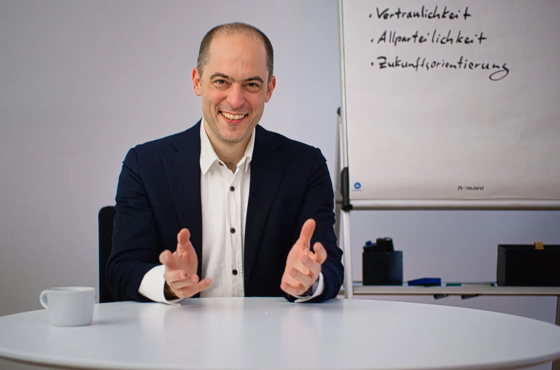 Christian von Baumbach at a table in a workshop room, behind him a flipchart with the words Vertraulichkeit, Allparteilichkeit and Zukunftsorientierung